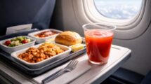 An airplane food meal tray on a fold-down table next to an aircraft window, featuring a plastic cup of bright red tomato juice.
