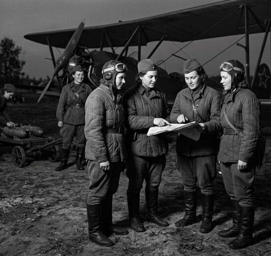 Soviet Night Witches female pilots and crew preparing for a night mission next to a Polikarpov Po-2 bomber. Four pilots are studying a map while a ground crew member pushes a bomb cart.