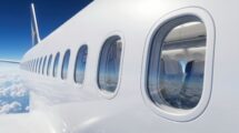 A close-up exterior view of a modern passenger jet featuring round airplane windows, with a blue sky and clouds in the background.